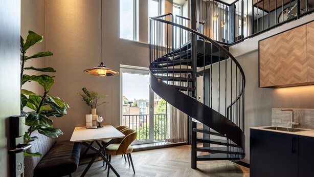 Une cuisine moderne avec un escalier en colimaçon noir, un coin repas avec un banc et des chaises, et de grandes fenêtres offrant une vue sur l’extérieur.