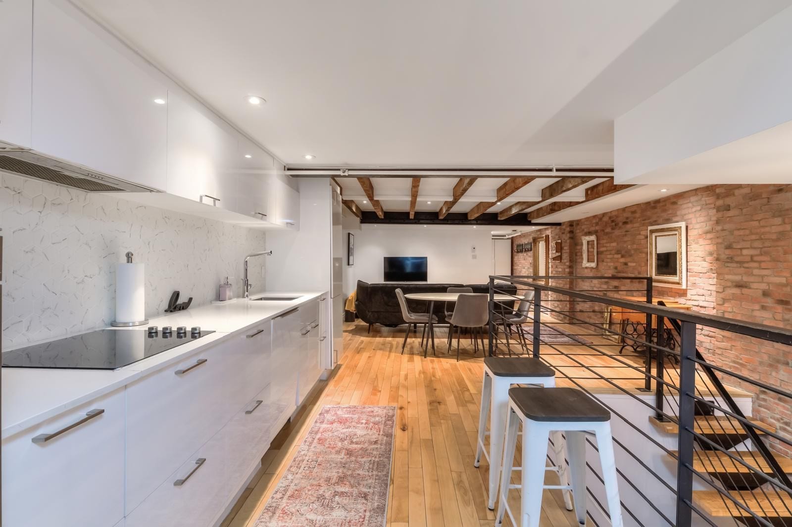 Eine moderne Küche mit weißen Schränken und einer zentralen Kochinsel, ein Essbereich mit Backsteinwand und eine Treppe mit Metallgeländer.