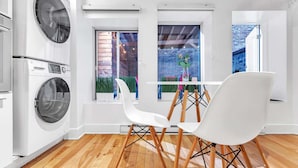 A modern kitchen with built-in appliances, a dining area with white chairs, and large windows offering views of a brick wall and greenery outside.