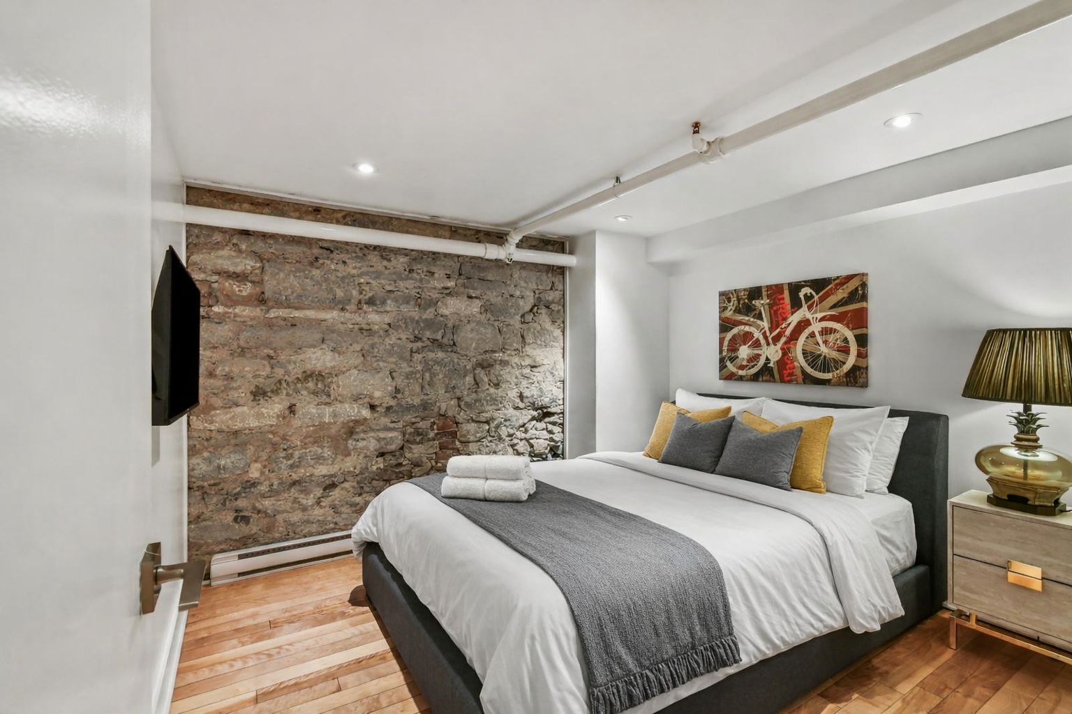 Une chambre moderne avec un mur en pierre, un lit agrémenté de plusieurs coussins, un téléviseur fixé au mur et un tableau accroché au mur.