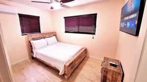 Un dormitorio con cama, cabecera de madera, ventilador de techo, dos ventanas con persianas, televisor en la pared y un pequeño armario de madera.