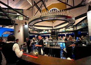 Zona de bar animada con una lámpara circular, estanterías llenas de botellas y gente interactuando.