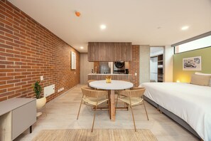 Ein modernes Hotelzimmer mit Backsteinwand, einer Essecke mit rundem Tisch und Stühlen, einem Bett mit weißer Bettwäsche und einer kleinen Küche im Hintergrund.