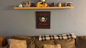Una sala de estar con un sofá marrón, cojines estampados, un peluche de Mickey Mouse y una estantería con varios objetos, incluyendo un casco de Star Wars y un póster.