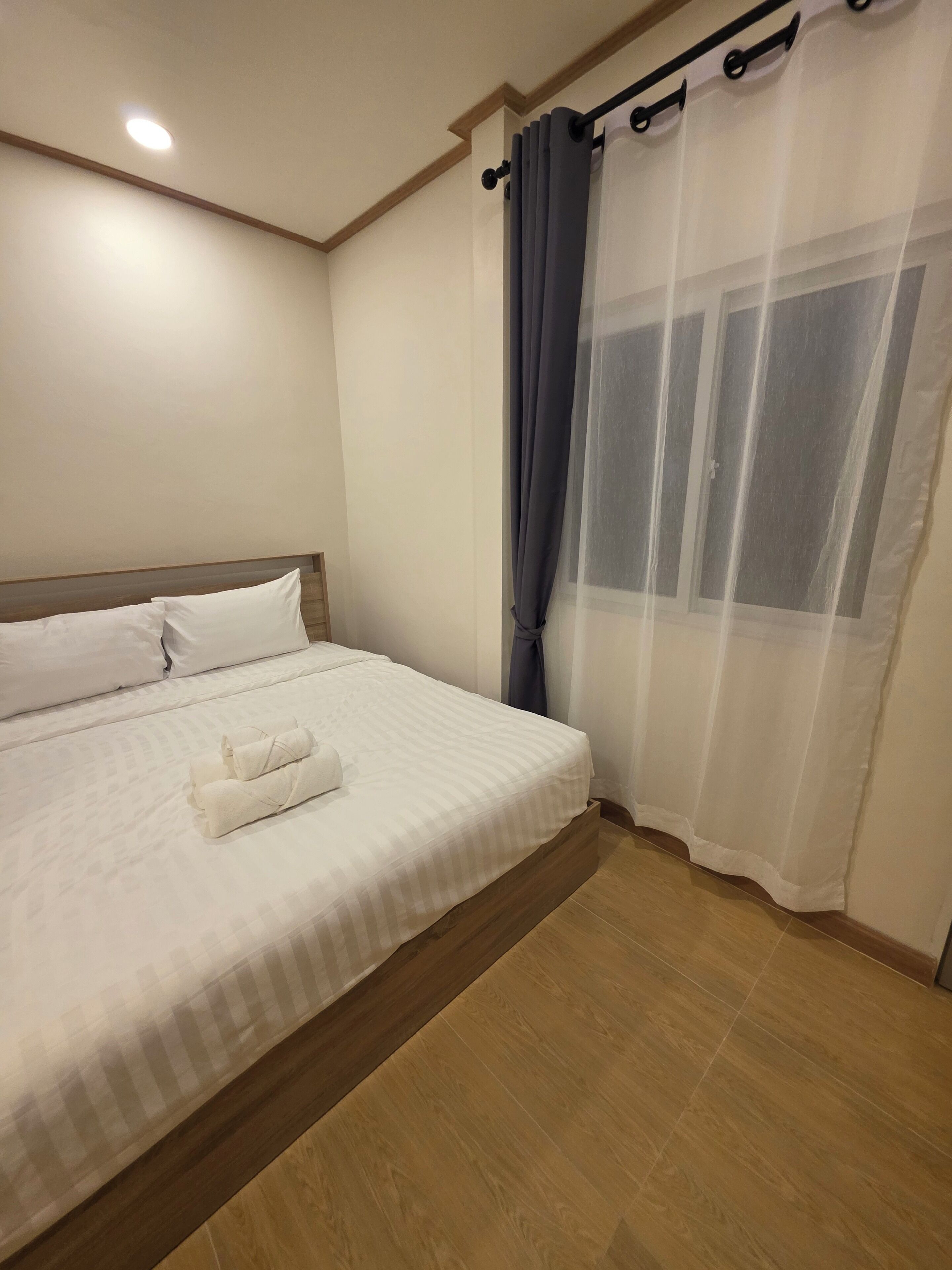 A neatly made bed with white linens and a folded towel, a wooden headboard, a ceiling light, and a window with sheer curtains.
