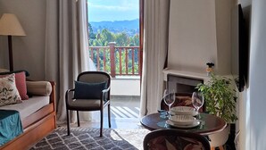 A living room with a sofa, armchair, and a table with wine glasses. A balcony with a railing and a view of greenery is visible through the window. - Duplex em Castelo Piscina e Lareira (Campos do Jordão)