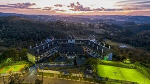 An aerial view of a large, multi-story building with a symmetrical facade, surrounded by lush greenery and a tennis court. - Duplex em Castelo Piscina e Lareira (Campos do Jordão)