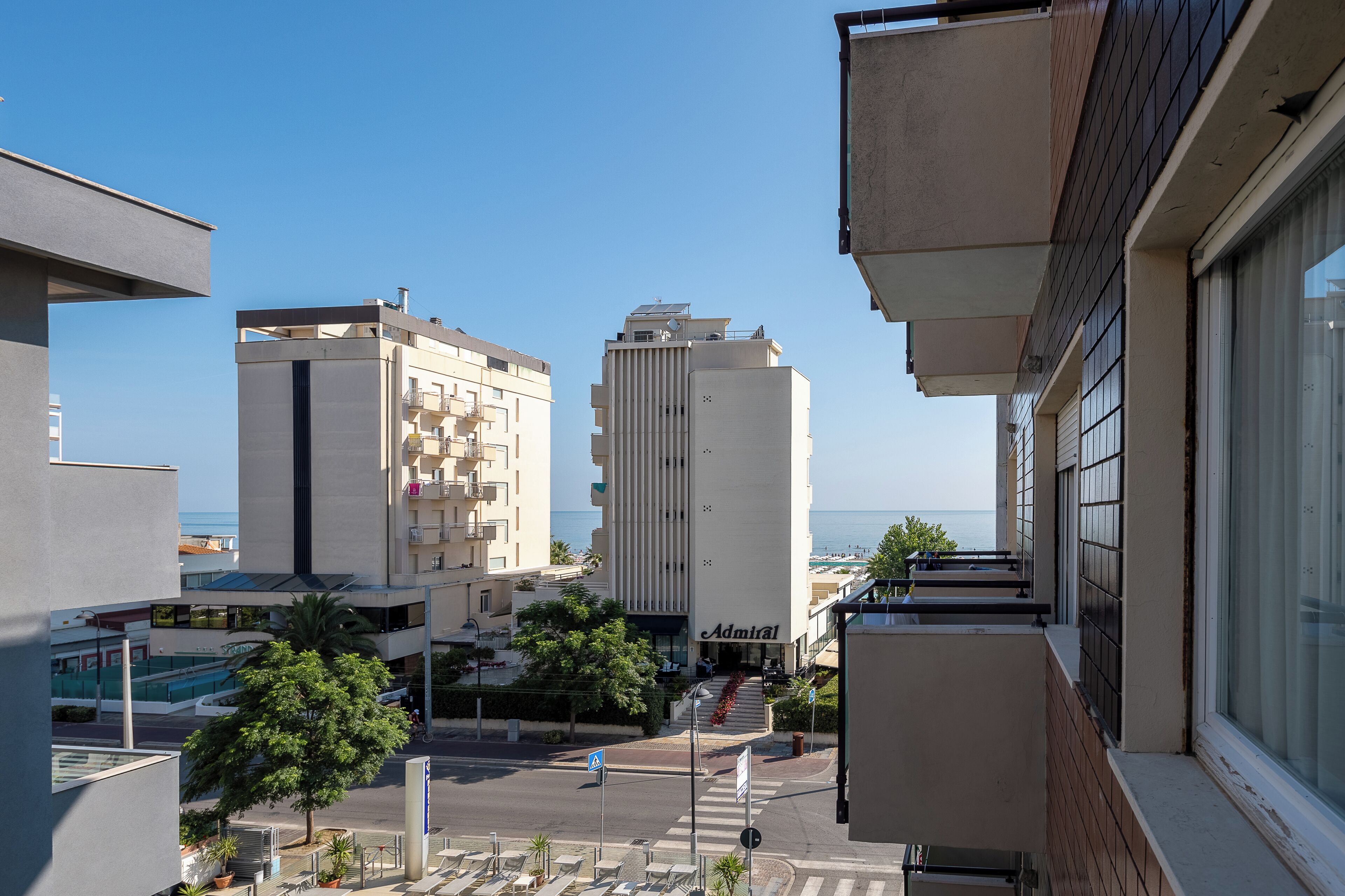 A modern building with a prominent 'Admiral' sign, a clear view of the sea, and a pedestrian area with trees and a crosswalk.