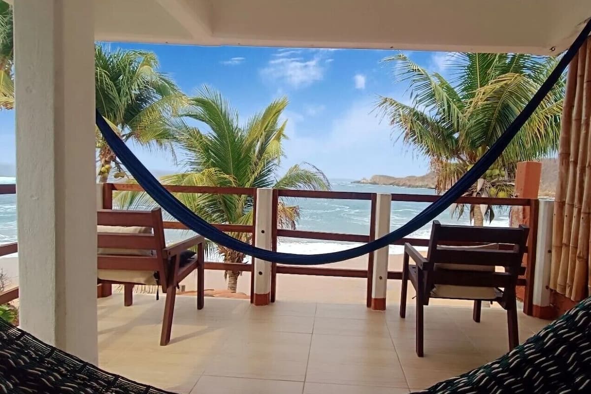 Un balcone con un'amaca, sedie in legno e vista sull'oceano e palme.