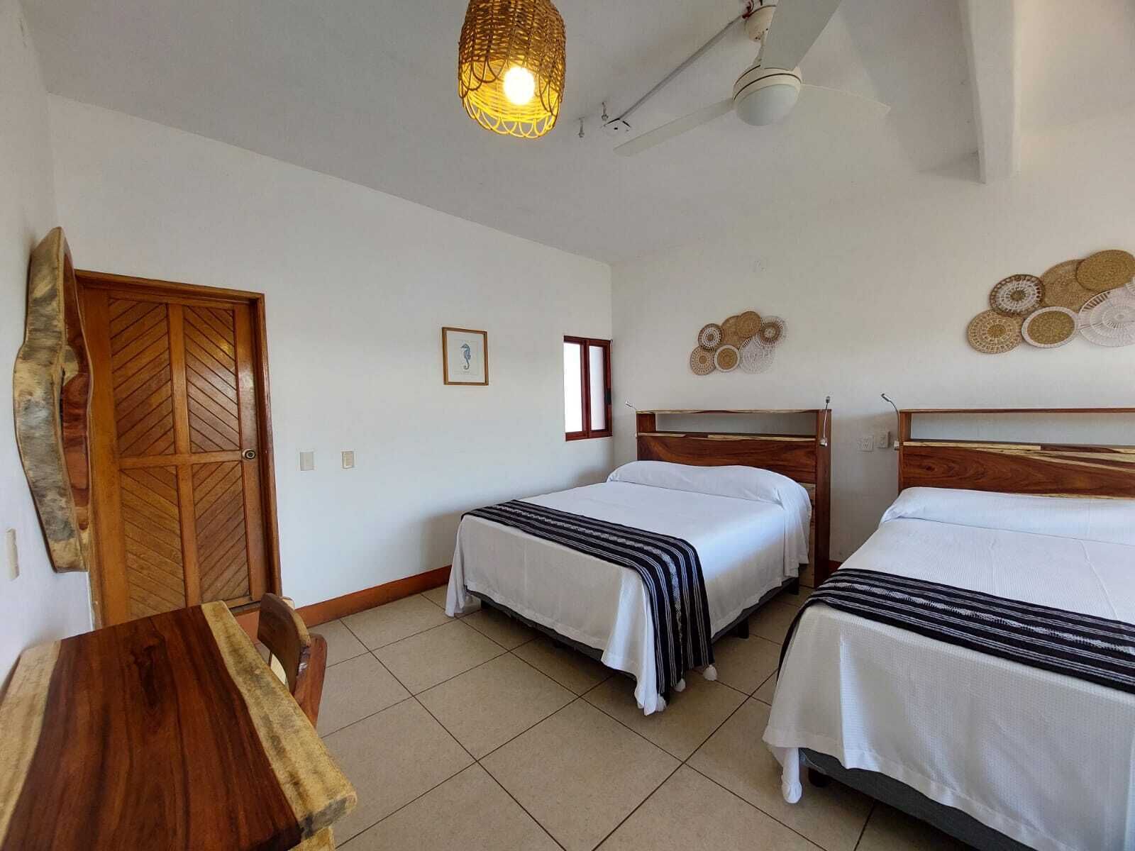 A hotel room with two beds, a wooden door, a woven light fixture, and a wall decoration.