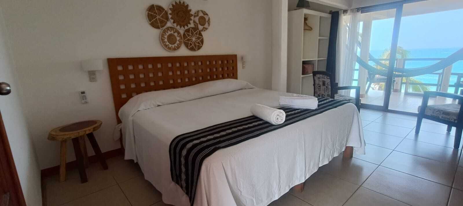 A hotel room with a bed, a small stool, a wall with decorative plates, and a view of the ocean through a glass door.