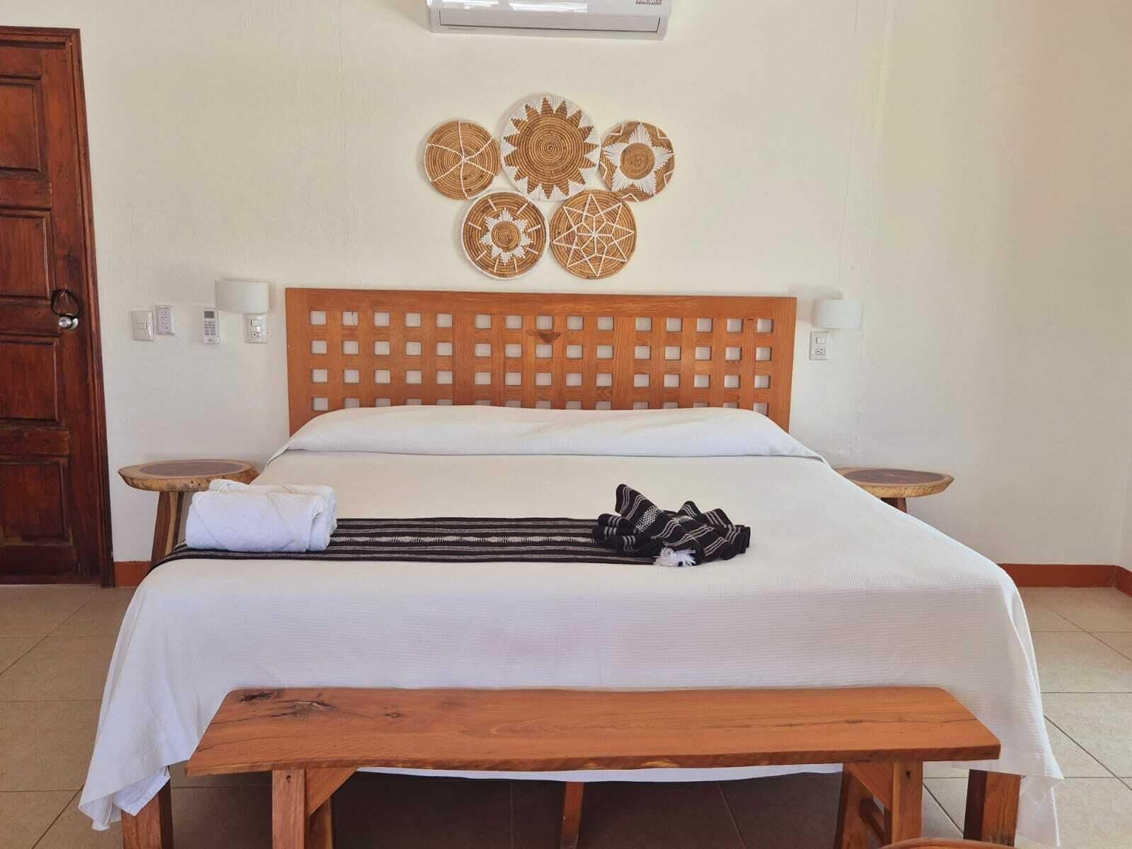 Une chambre d’hôtel avec un lit en bois, du linge de lit blanc, une tête de lit en bois, un panneau décoratif fixé au mur, des lampes de chevet et un banc en bois.