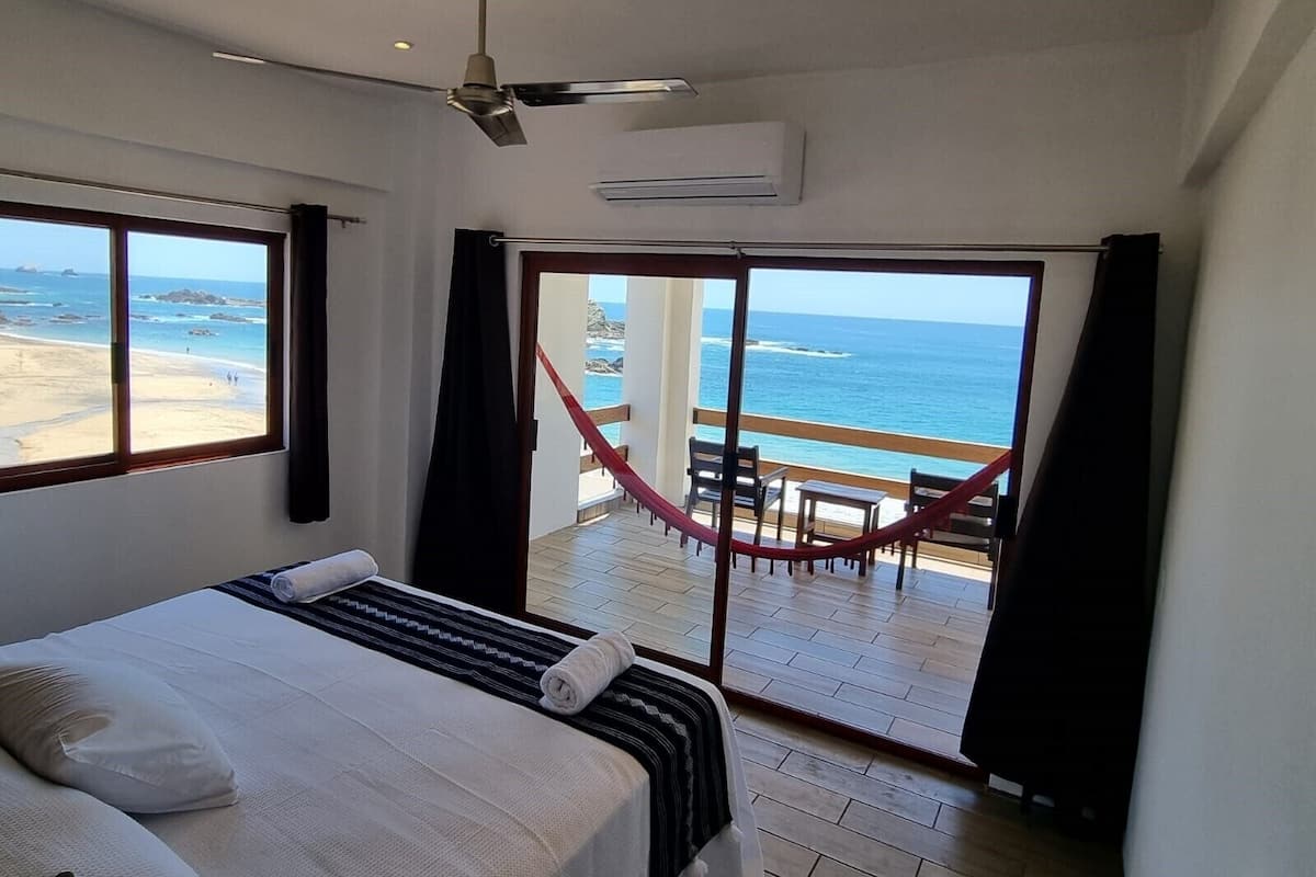 Una camera d'albergo con un letto, una ventola a soffitto, un condizionatore d'aria e vista sulla spiaggia attraverso un'ampia finestra.