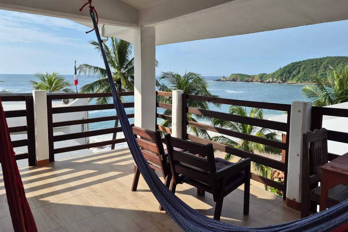 Un balcone con un'amaca, sedie di legno e vista sull'oceano e sulle colline.