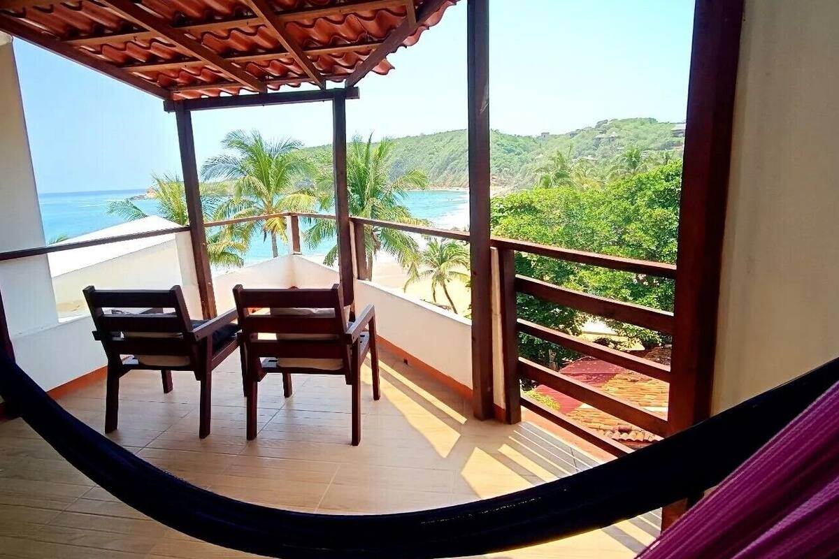 Un balcone con due sedie di legno e un'amaca, con vista su una spiaggia e vegetazione lussureggiante.