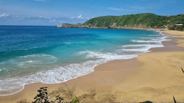 Ein Strand mit klarem türkisblauem Wasser, sandigem Ufer und einer bewaldeten Hügelkette im Hintergrund.