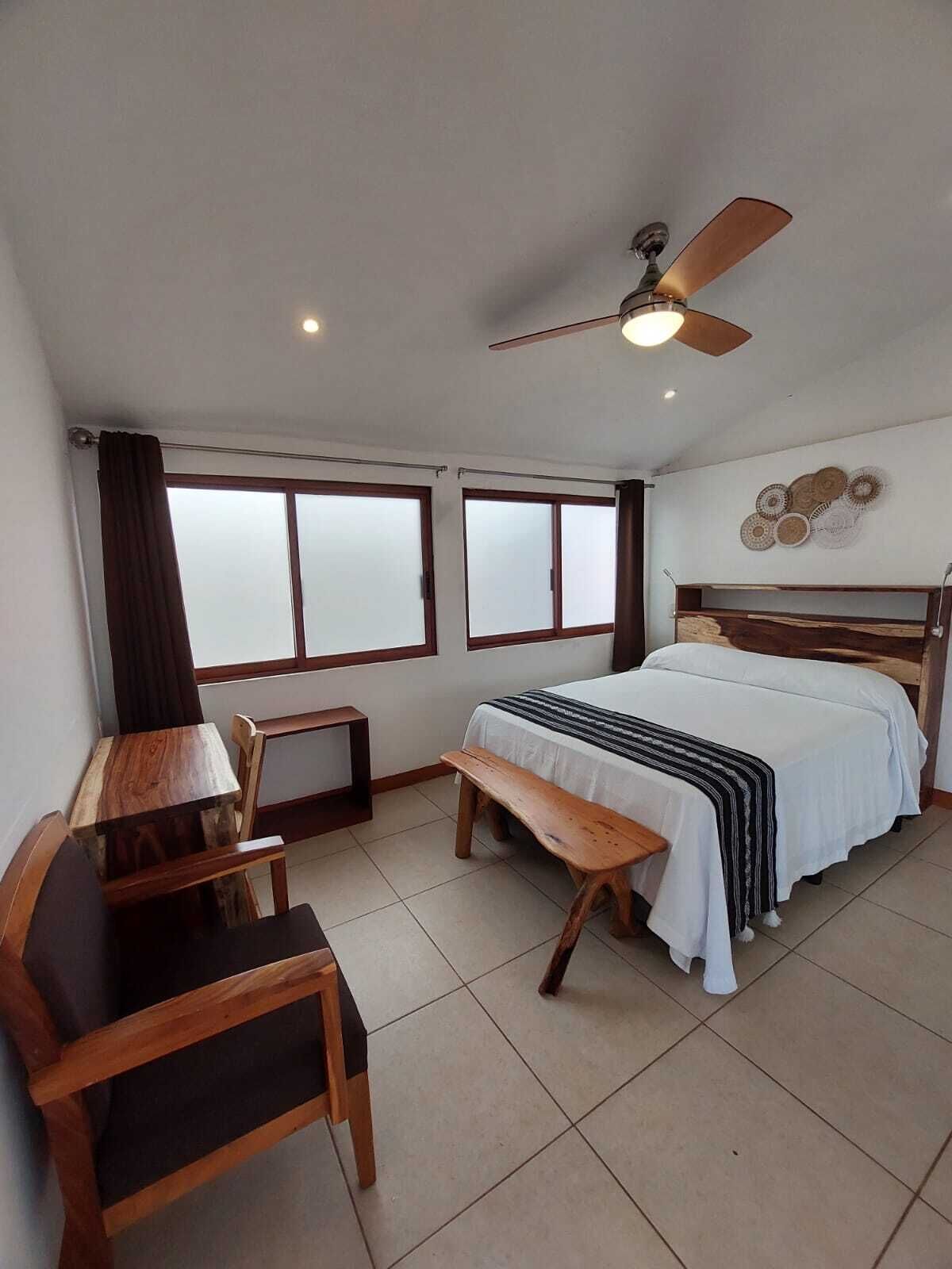A bedroom with a bed, a desk, a chair, a ceiling fan, and large windows.