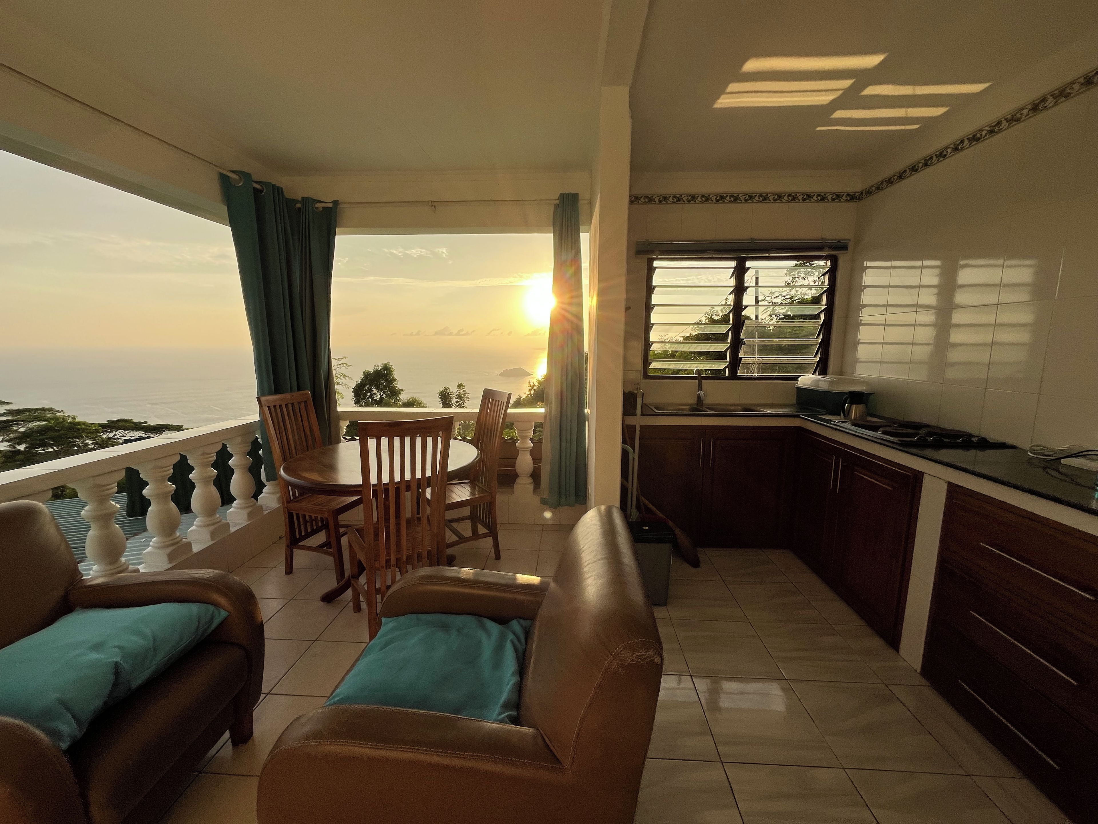 A balcony with a table and chairs, a sofa, a kitchen area with a sink and stove, and a view of the sea.