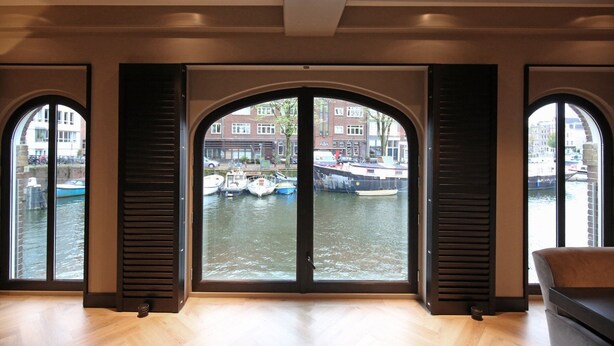 A room with large windows overlooking a canal with boats, featuring a modern interior with wooden flooring and dark shutters. - Luxury, Newly Remodeled Apartment in 16th Century Warehouse, with 10 meter Boat. (amsterdam)