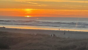 A beach with the sun setting over the horizon, waves gently crashing onto the shore, and a few people walking along the beach.