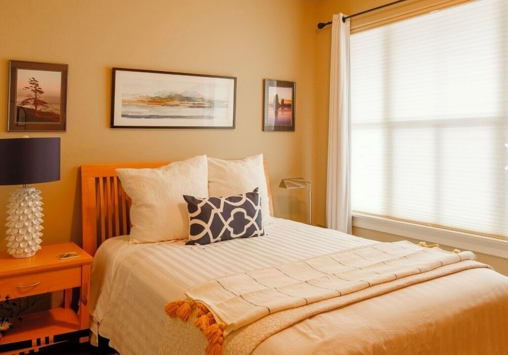 A neatly arranged bedroom with a bed, a bedside table with a lamp, and framed pictures on the wall.
