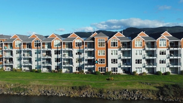A row of modern multi-story residential buildings with balconies, situated beside a body of water with a mountainous backdrop.
