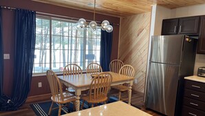 Une cuisine aux murs lambrés, dotée d’un réfrigérateur en acier inoxydable, d’une table à manger en bois avec des chaises, et d’une fenêtre agrémentée de rideaux.