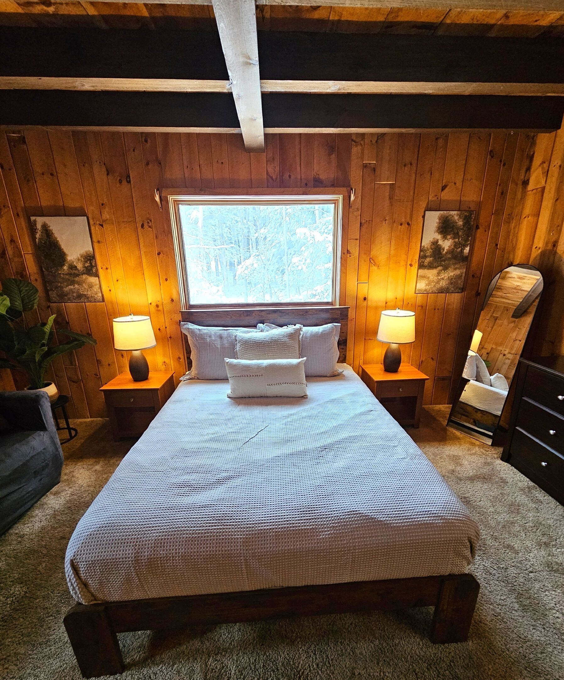 Una camera da letto accogliente con soffitto in legno, un letto grande con copriletto decorato, due lampade sui comodini e una finestra con vista sugli alberi.