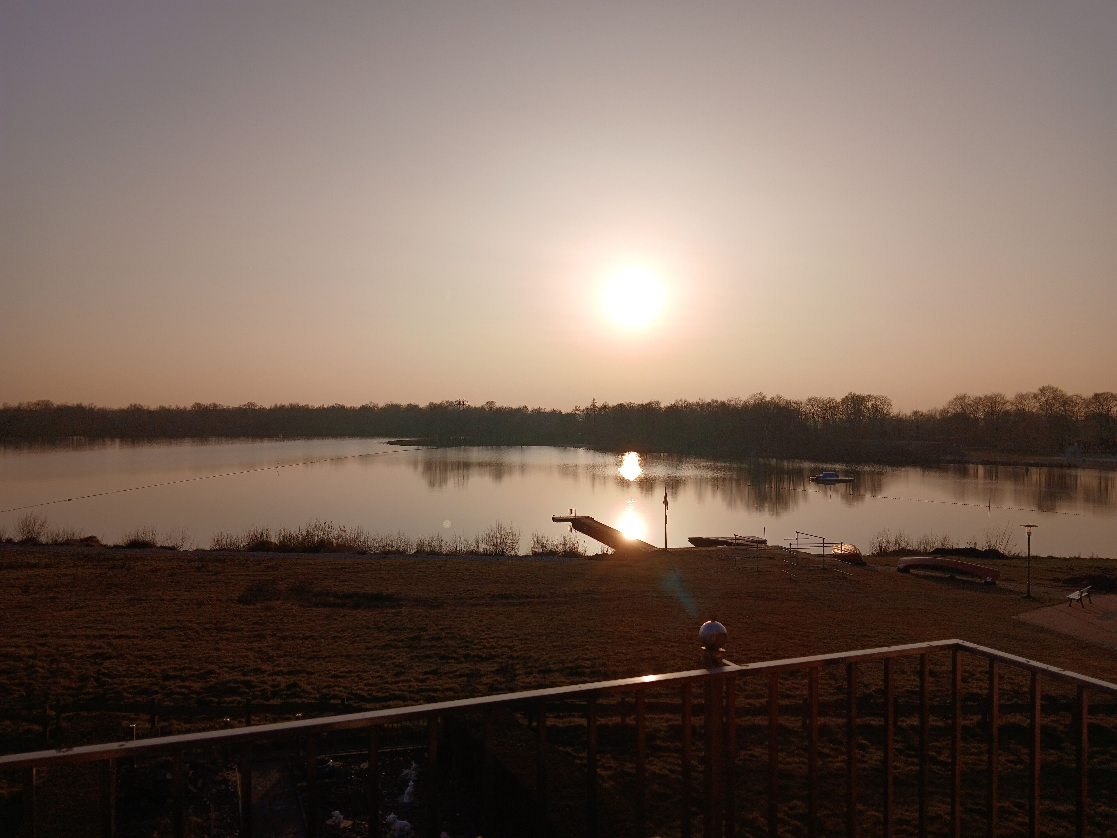 Ein ruhiger See bei Sonnenuntergang mit blauem Himmel, einer Anlegestelle und einer fernen Küste.