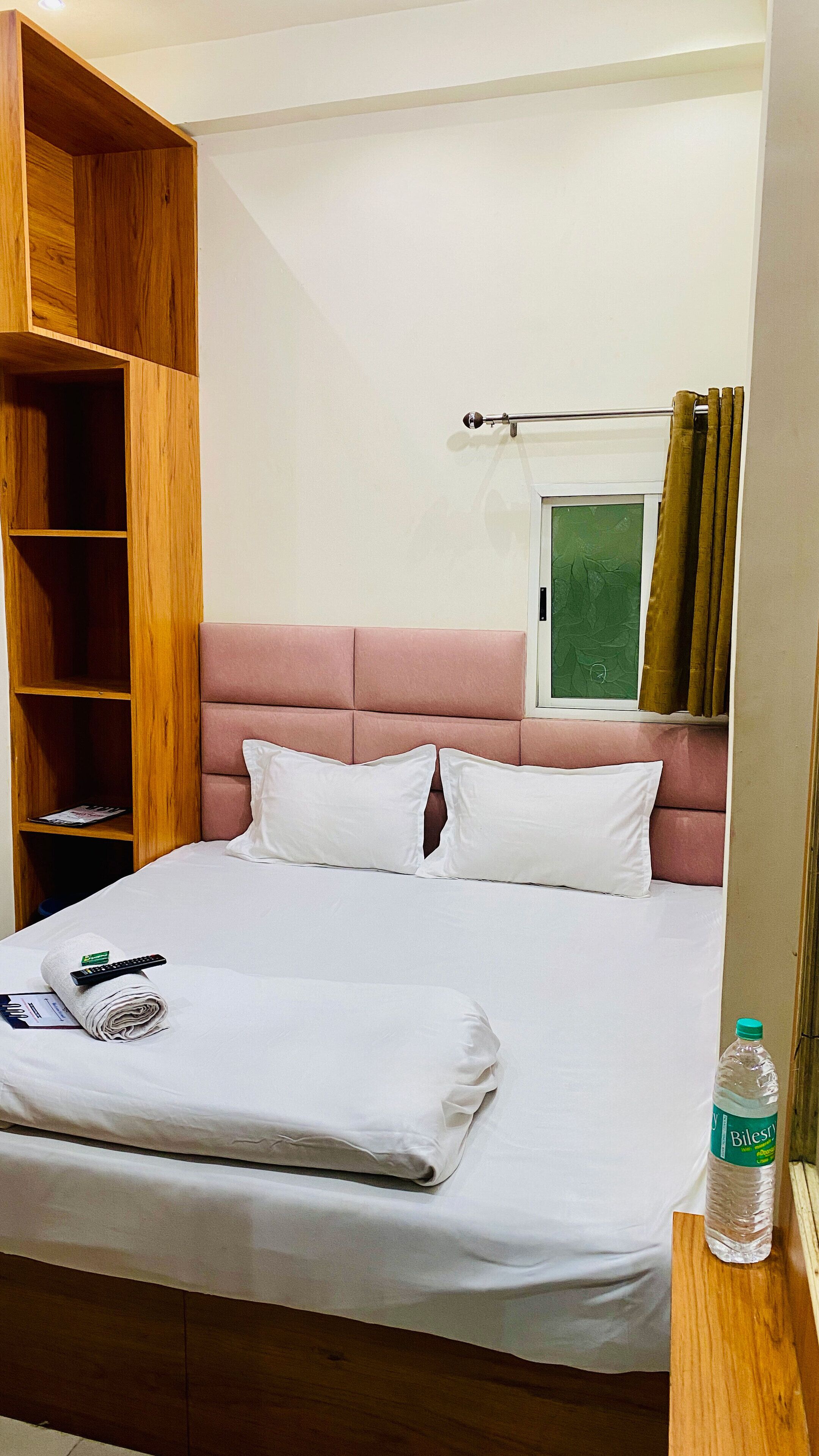 A neatly made bed with pillows, a wooden wardrobe, a window with curtains, and a bottle of water on a wooden nightstand.