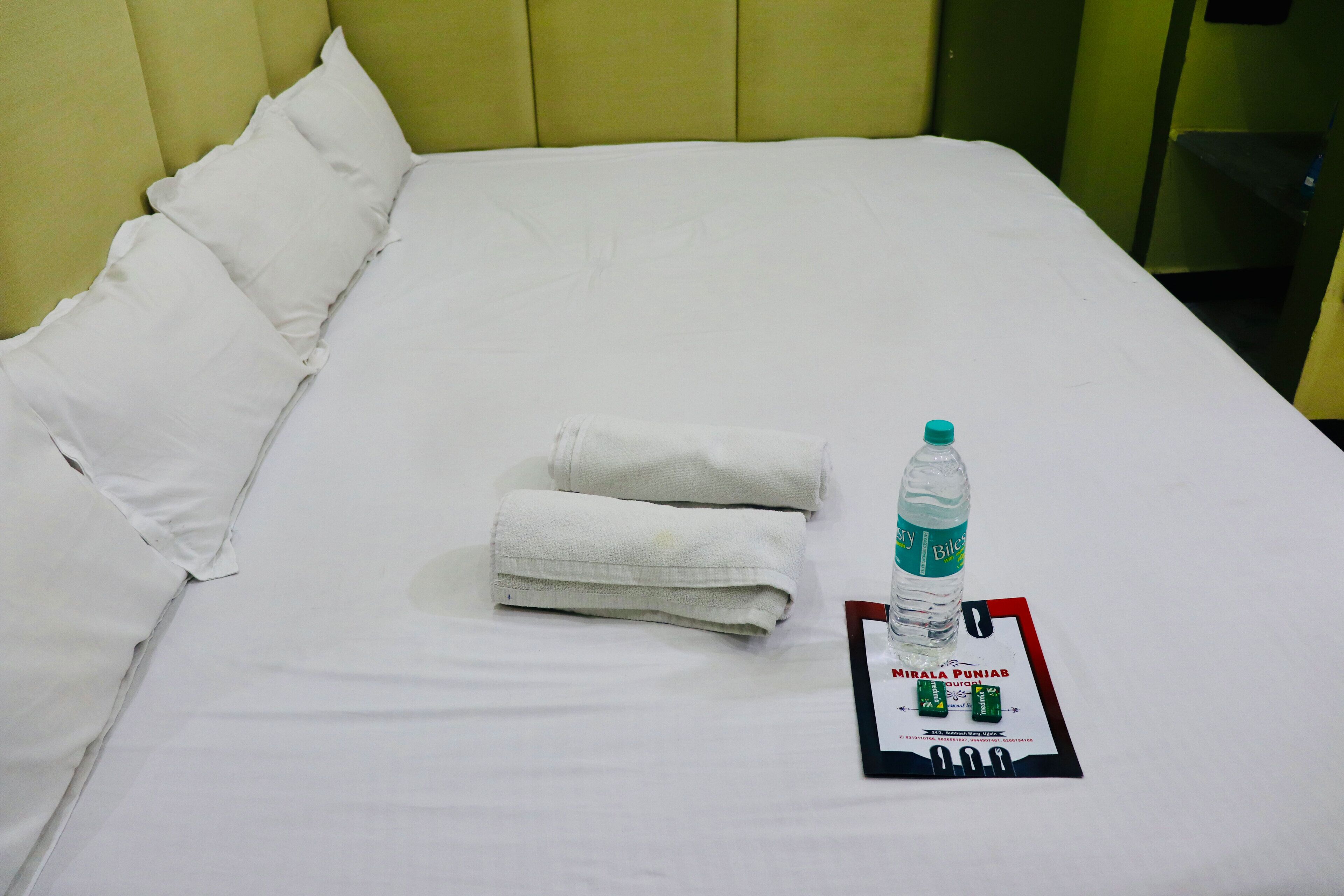 A neatly made bed with white linens, a folded towel, a bottle of water, and a card with the text "Mirala Punjab."