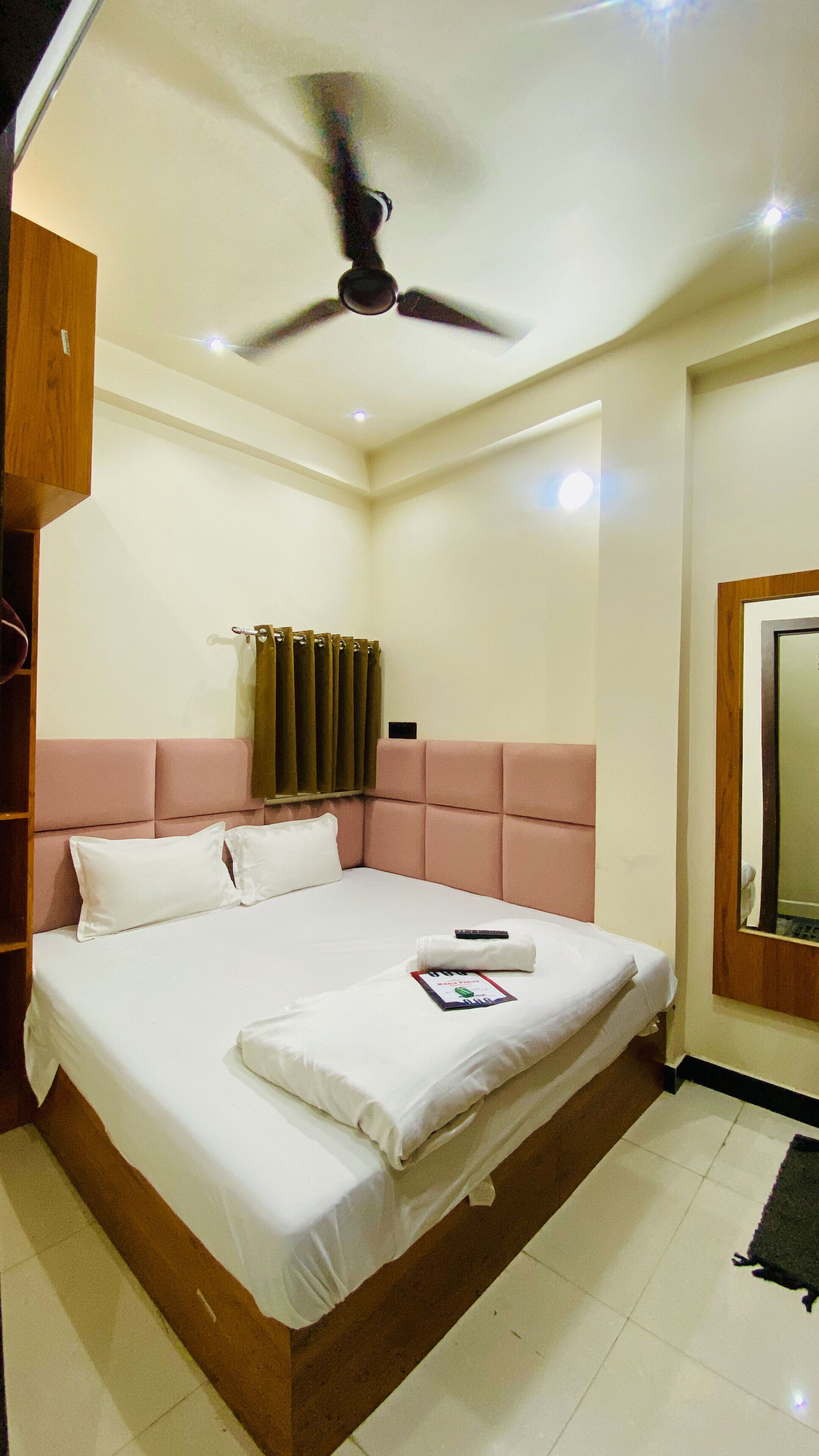 A neatly made bed with white linens and pillows, a ceiling fan, a window with curtains, and a wooden headboard.