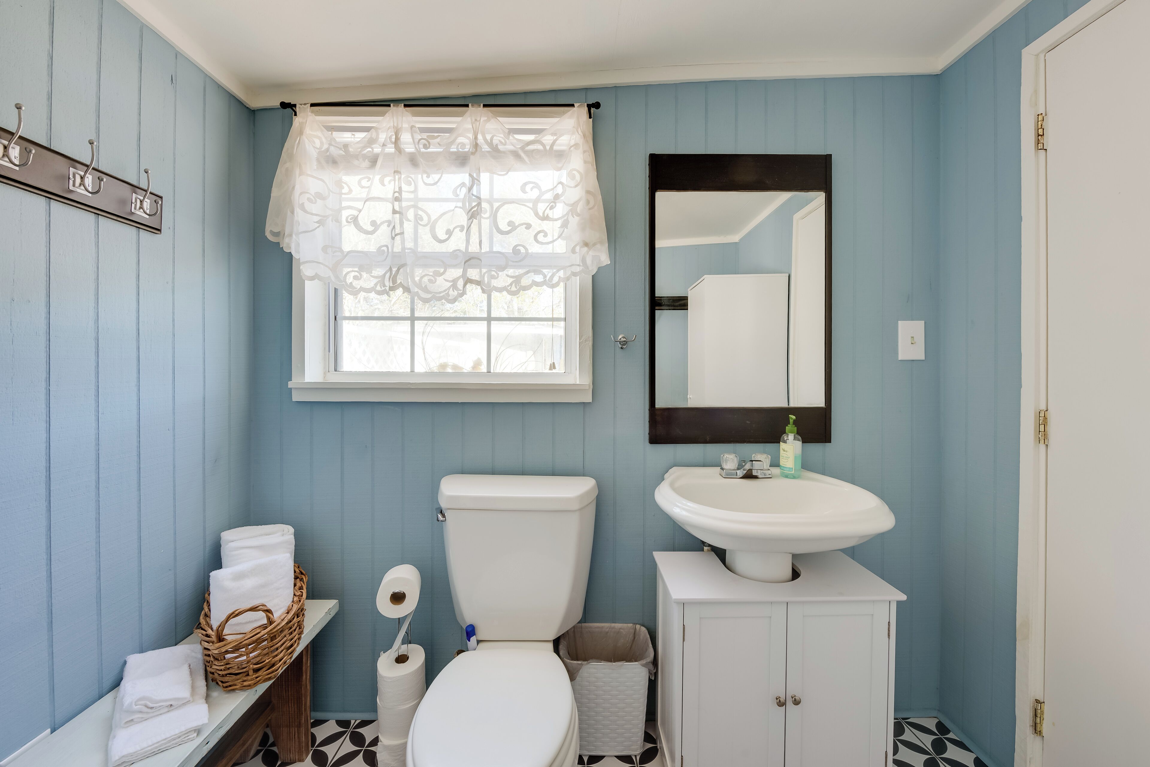 A bathroom with a toilet, sink, and a mirror. There is a window with a curtain, a basket of towels, and a roll of toilet paper.