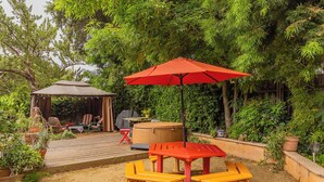 Terraza de madera con un paraguas rojo, una mesa y sillas. Hay una zona de estar cubierta con una parrilla y luces colgantes.