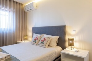 A hotel room with a bed, two bedside tables, a wall-mounted air conditioner, and a window with curtains. - Beachouse Pvz 🌊🌅 (Póvoa de Varzim)