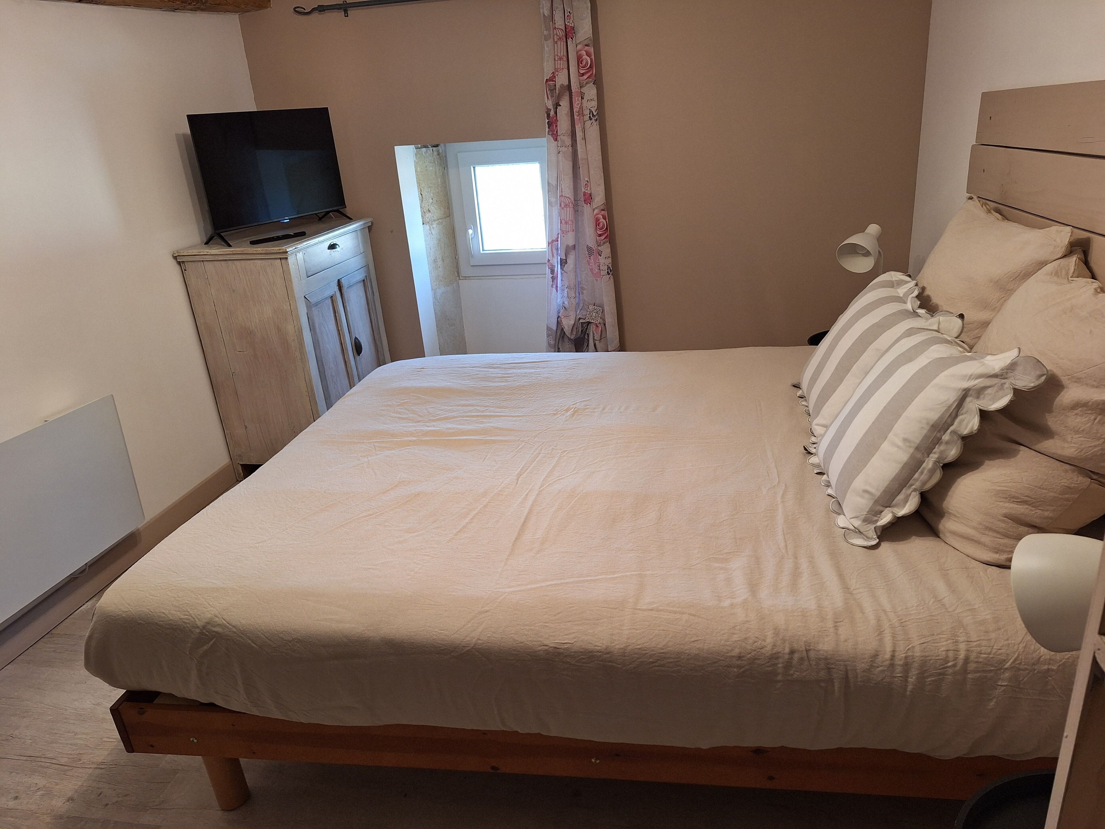 Une chambre à coucher avec un lit, un téléviseur posé sur un meuble, une fenêtre avec des rideaux et une lampe de chevet.