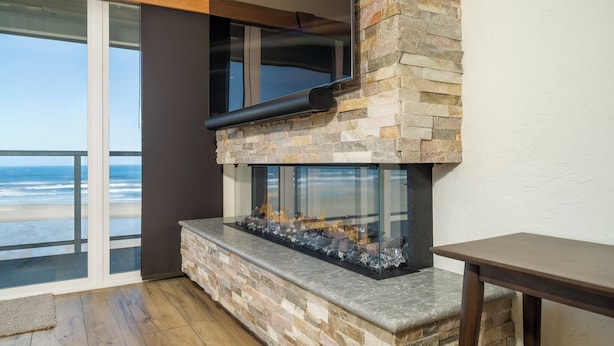 A modern living room with a stone fireplace, large windows overlooking the beach, and a wooden ceiling. - Experience Nature's Spectacle from the Front Row! (Newport)