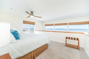 A bedroom with a bed, a ceiling fan, a window with a view of the beach, and a wooden bench.