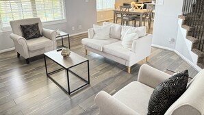 A modern living room with a beige sofa, a matching armchair, and a coffee table. There is a staircase to the right and a kitchen in the background.
