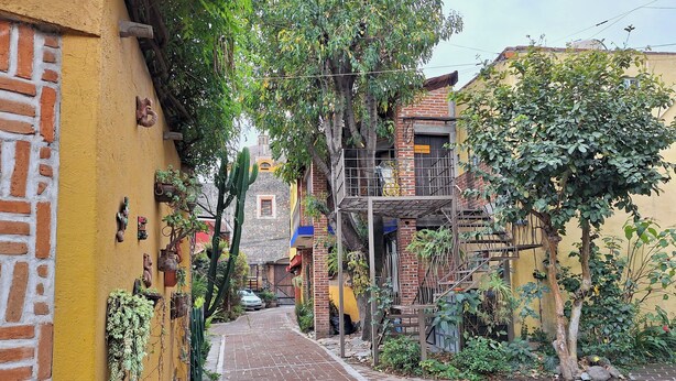 Une ruelle étroite en briques, avec un immeuble jaune sur la gauche et un immeuble à plusieurs étages doté d’un escalier métallique sur la droite.