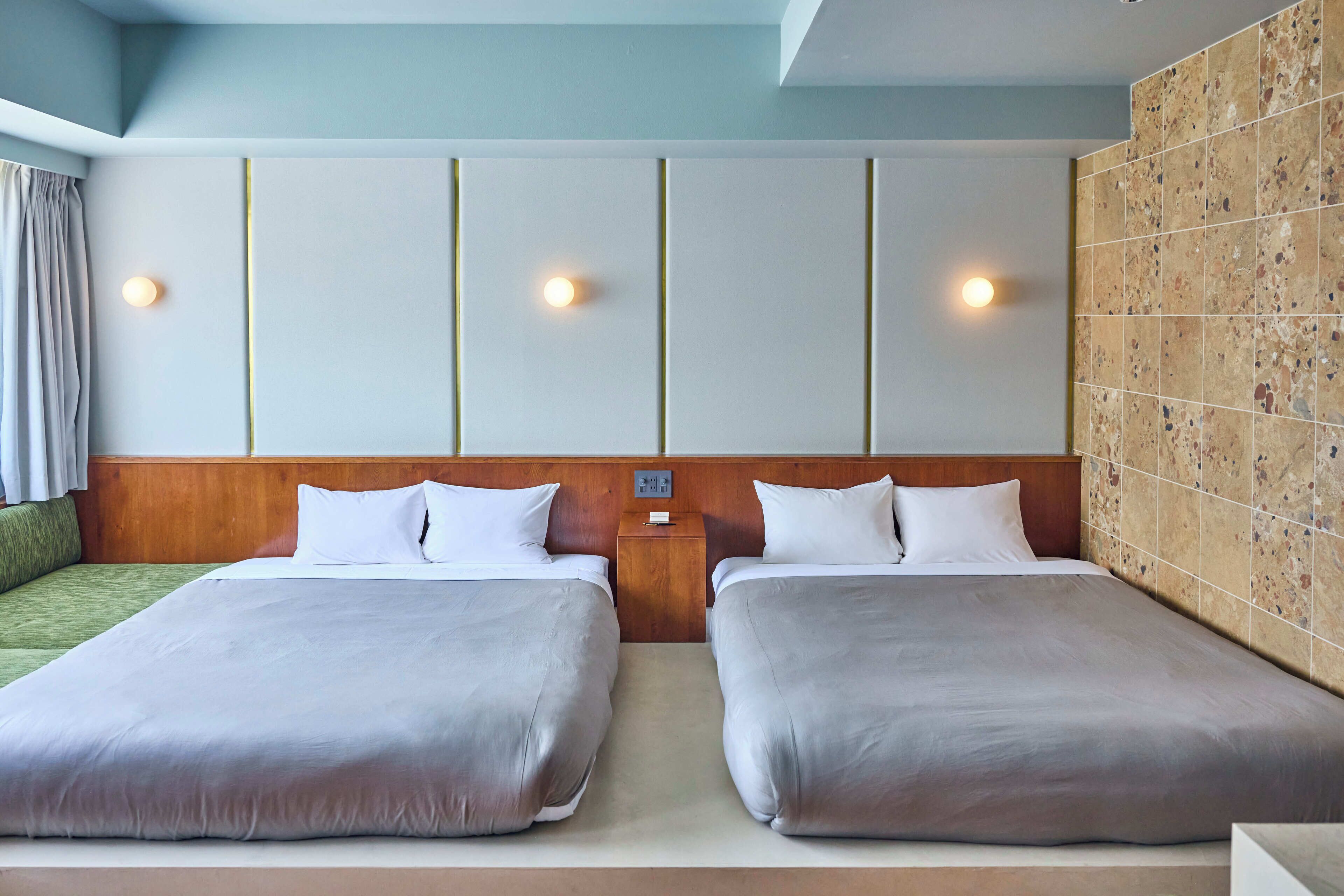 Ein Hotelzimmer mit zwei Betten, einem hölzernen Kopfteil, einem Nachttisch und einer Wand mit strukturierter Fliesenoptik.