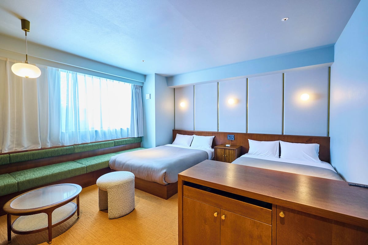 A hotel room with a large bed, a seating area with a bench and a round table, a wooden dresser, and a window with curtains.