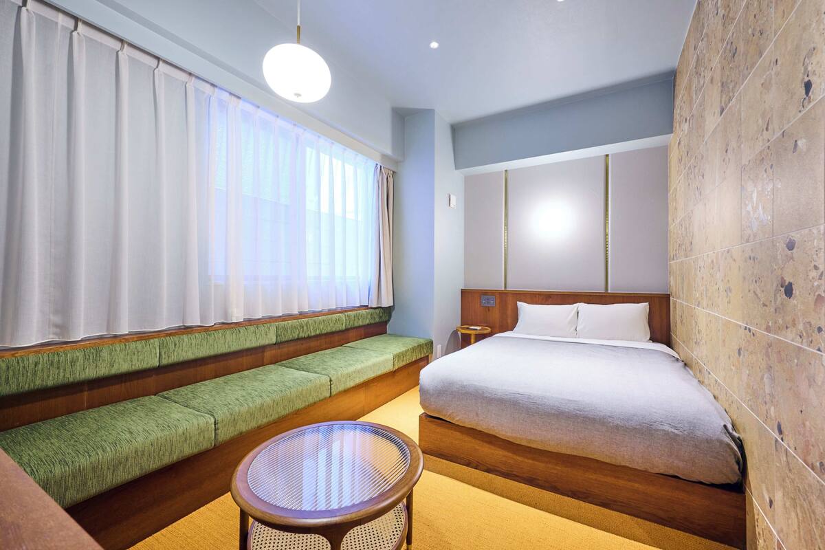 A hotel room with a large bed, a green cushioned bench, a round table, and a large window with sheer curtains.