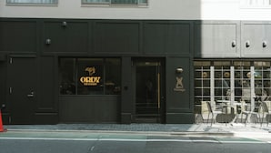 Un edificio con fachada oscura, un escaparate visible con el letrero "ORDY THE BAKERY" y una sección con mesas al aire libre.