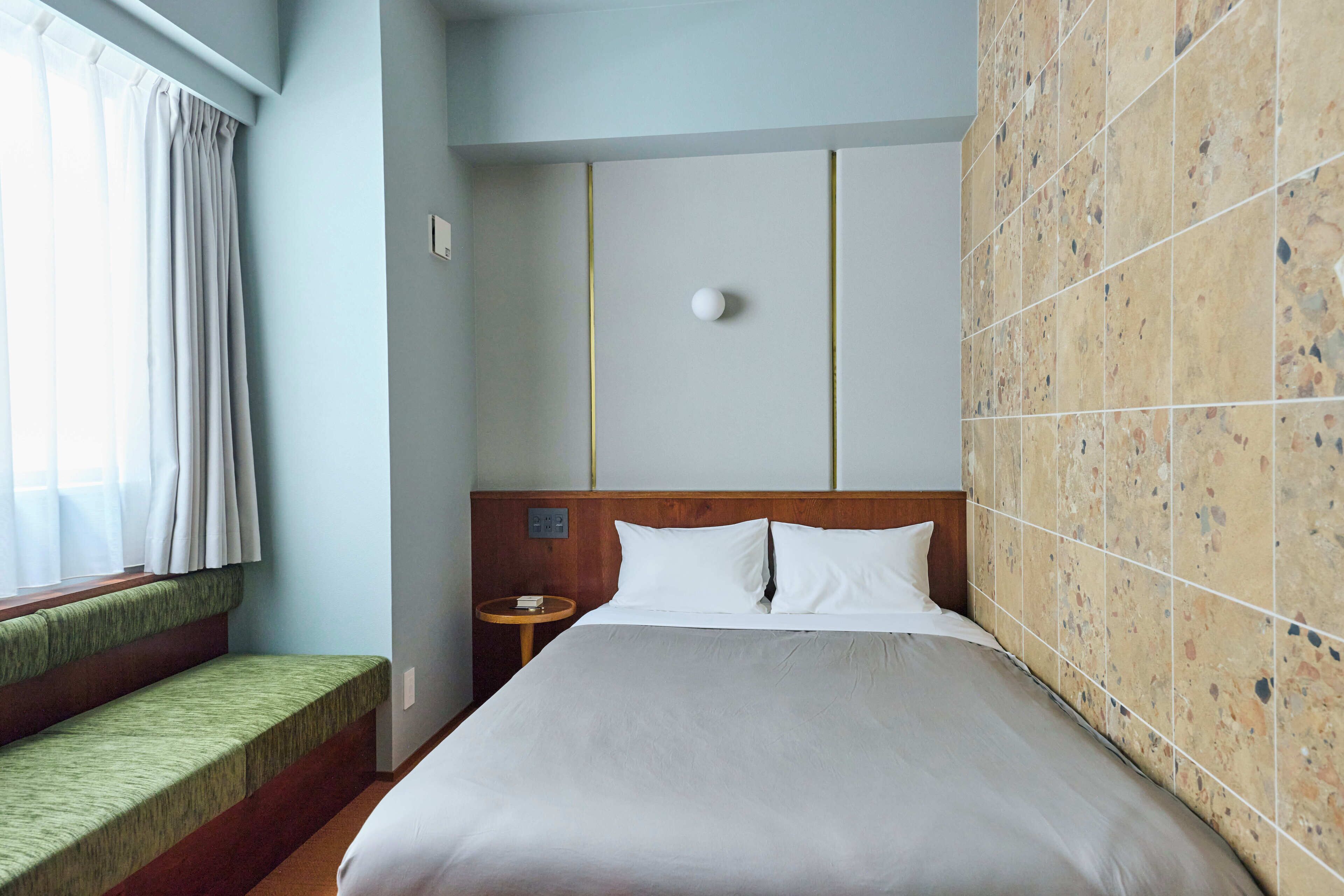 Ett hotellrum med en säng, ett litet sängbord, en bänk vid fönstret och en vägg täckt med kakel.