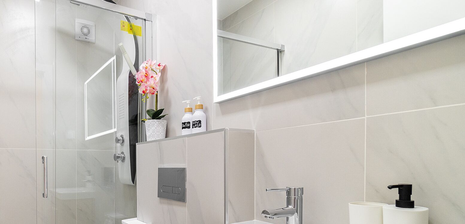 Un bagno moderno con box doccia in vetro, lavandino bianco e specchio sopra il lavandino. Sul piano del lavandino ci sono due flaconi di bagnoschiuma.