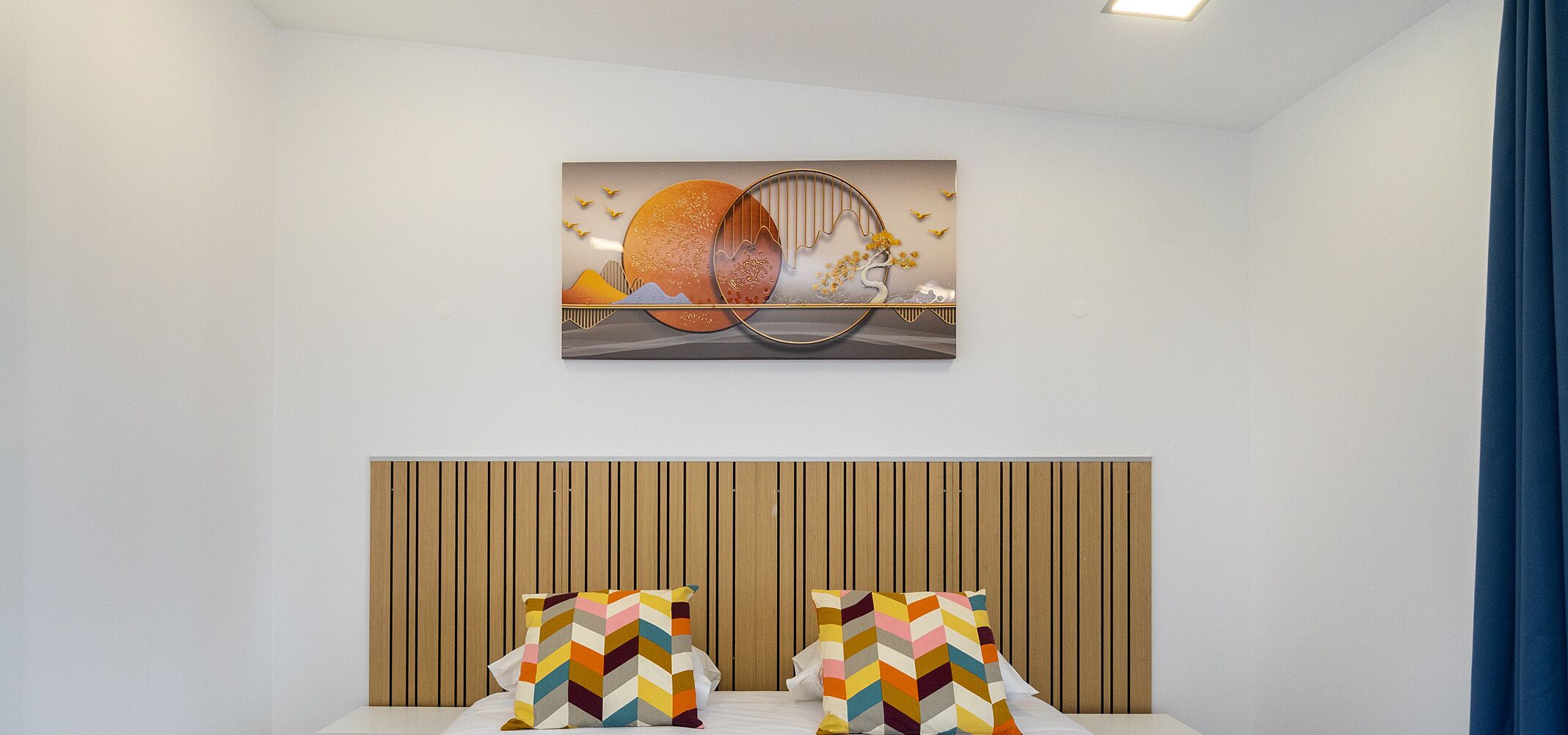 A bedroom with a wooden headboard, two colorful pillows, and a wall-mounted artwork depicting a sunset and birds.