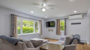 A modern living room with a ceiling fan, large windows, a flat-screen TV, and a wooden table. - 3-bedroom Cozy Home in Ellsworth with AC, WiFi (Ellsworth)