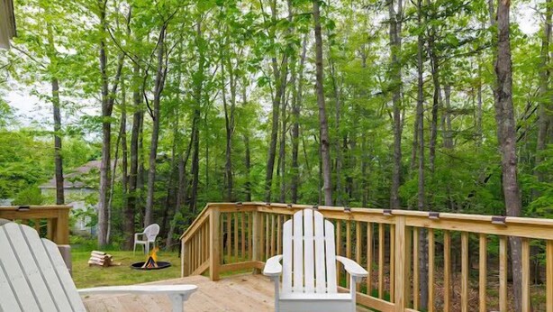 A wooden deck with white Adirondack chairs and a glass-top table, surrounded by a forest. - 3-bedroom Cozy Home in Ellsworth with AC, WiFi (Ellsworth)