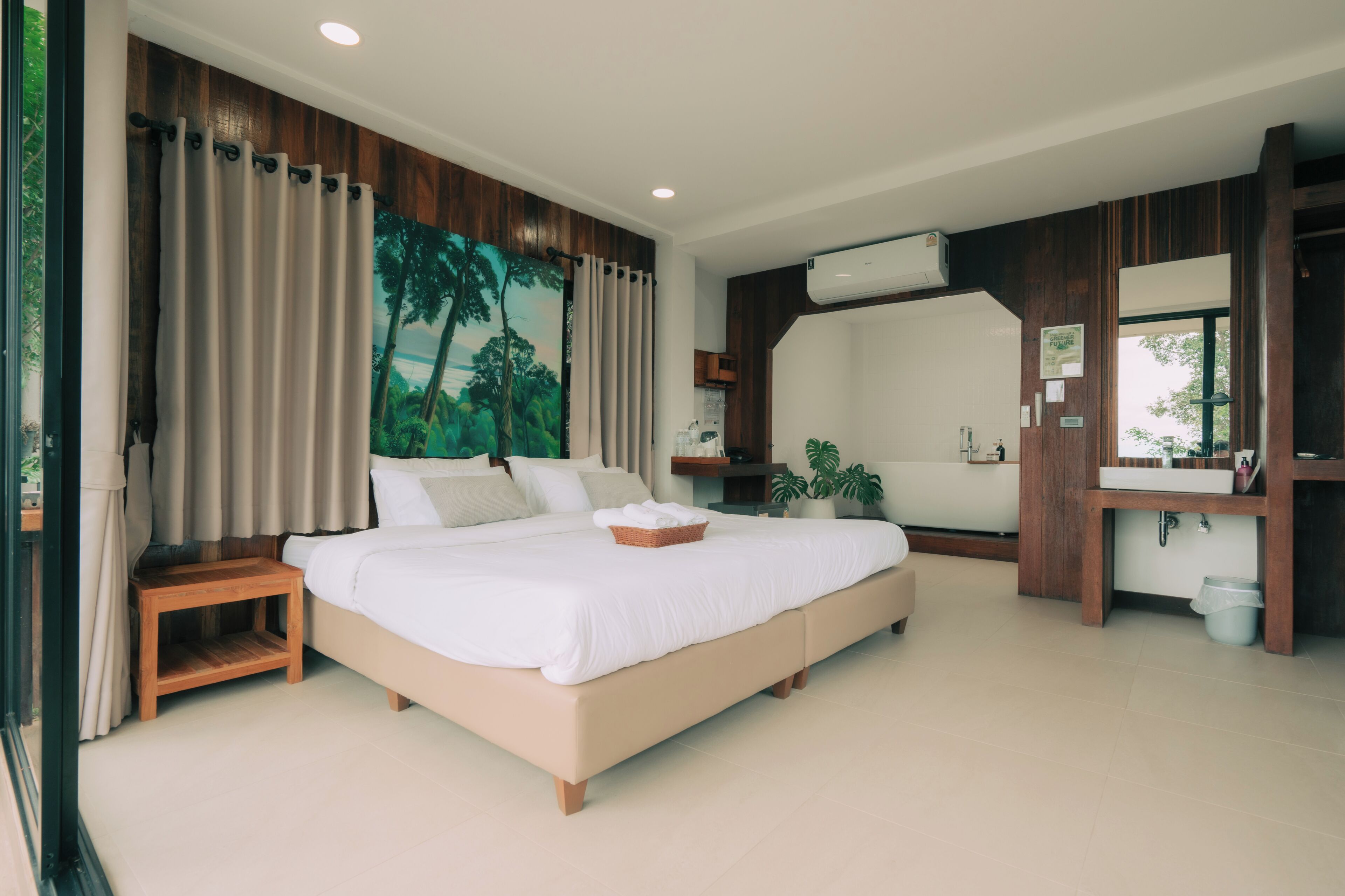 A bedroom with a large bed, a wooden nightstand, a wall-mounted air conditioner, a bathroom with a sink and mirror, and a large window with curtains.